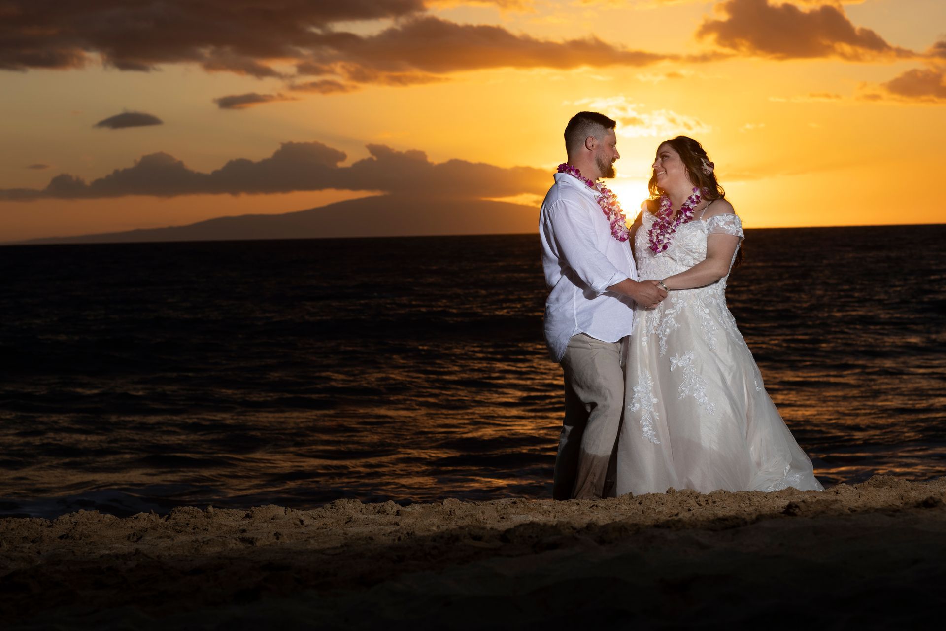 Couple holding hands on a beach at sunset, wearing leis.