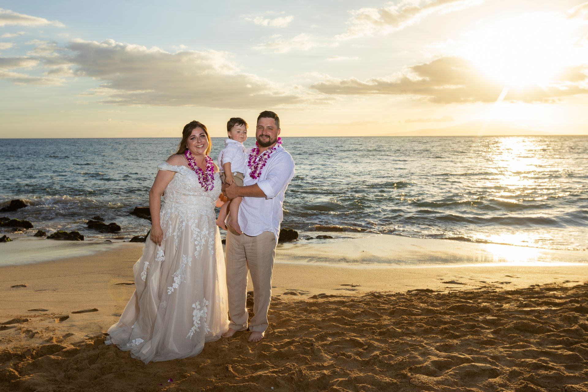 Family on beach at sunset, wedding dress, holding child, all wearing leis.