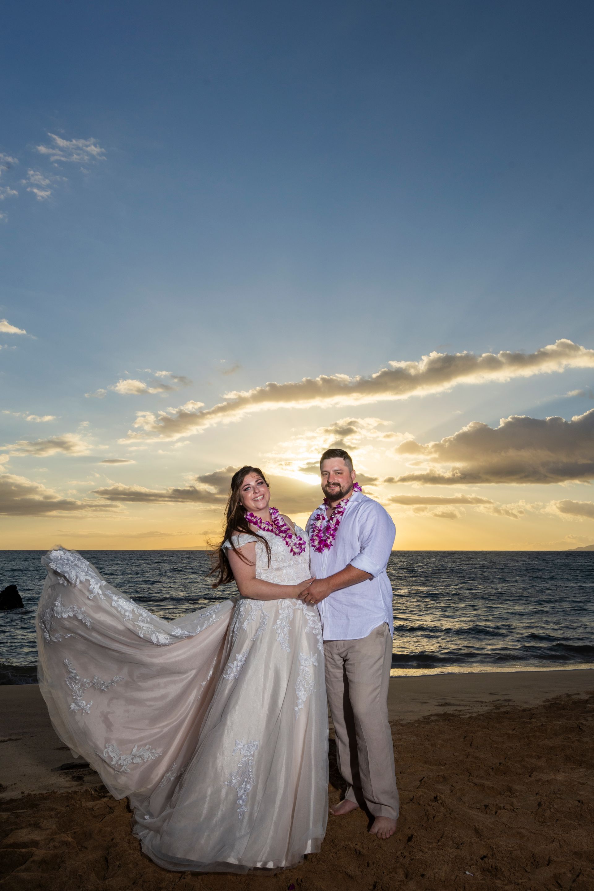 Couple on beach at sunset, woman in flowing gown, both wearing leis, holding hands.