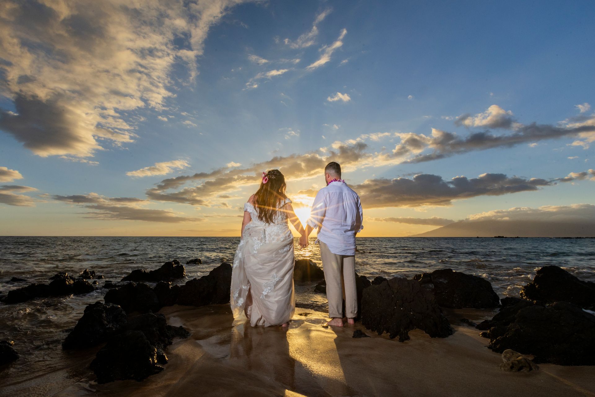 Newlyweds holding hands on a beach, facing sunset. Warm colors, water, rocks, and cloudy sky.