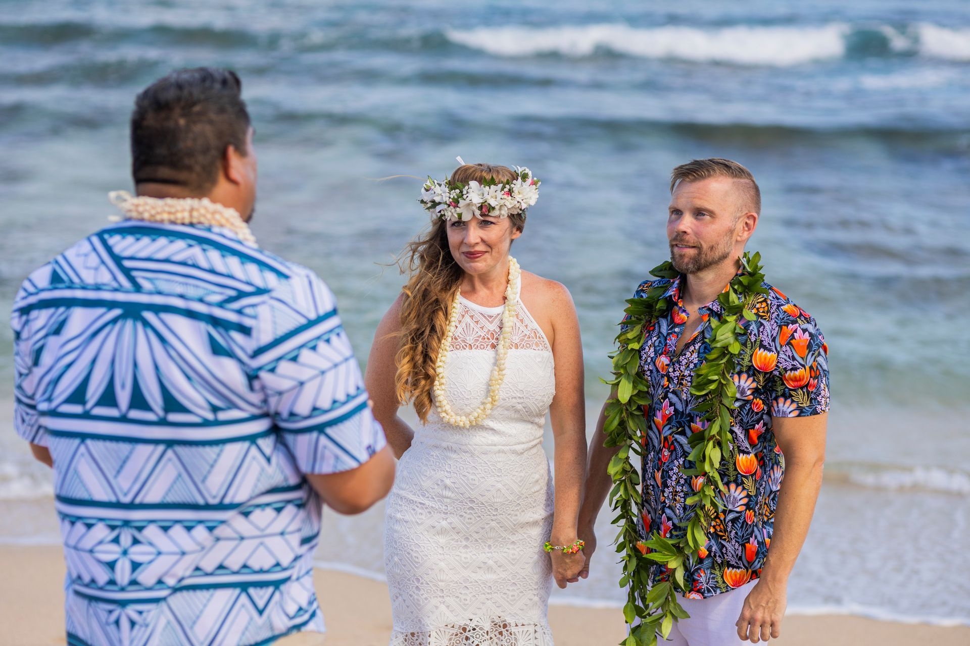 A couple holds hands during a beach wedding ceremony, officiated by a man in a patterned shirt.