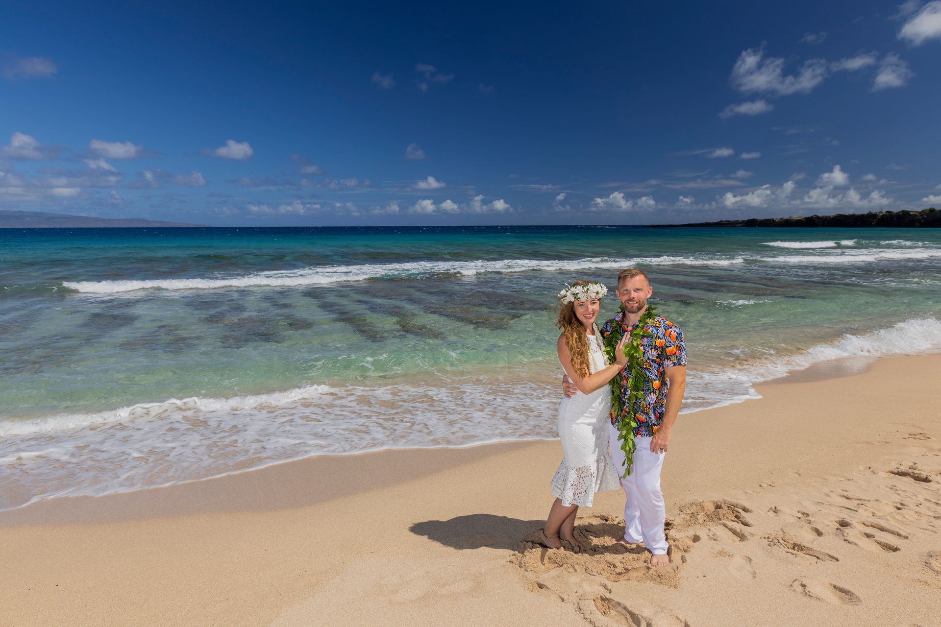 Couple on beach, bride in white dress, groom in floral shirt, blue ocean, sunny day.