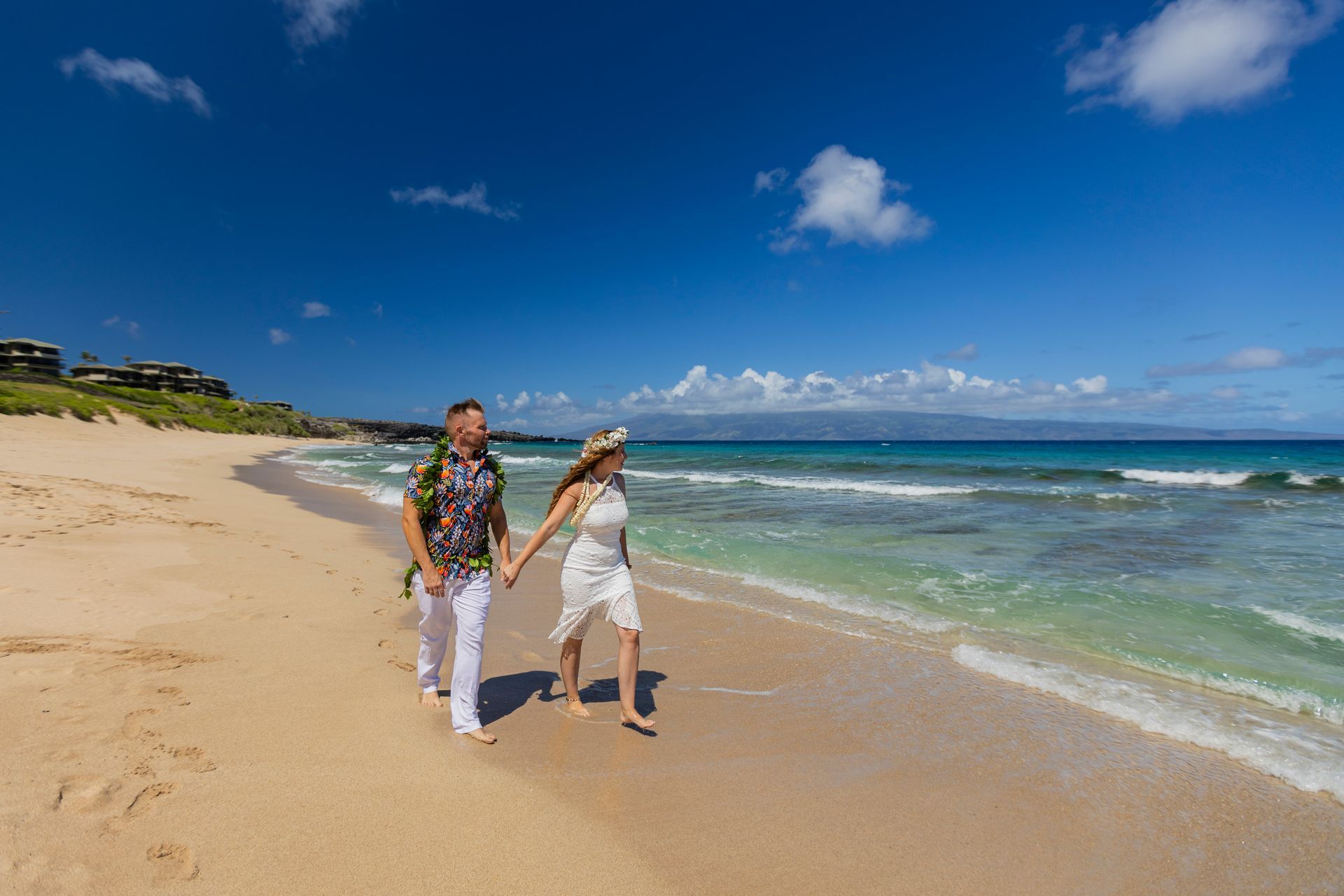 Couple walks along a sandy beach, holding hands; clear blue sky and ocean in background.
