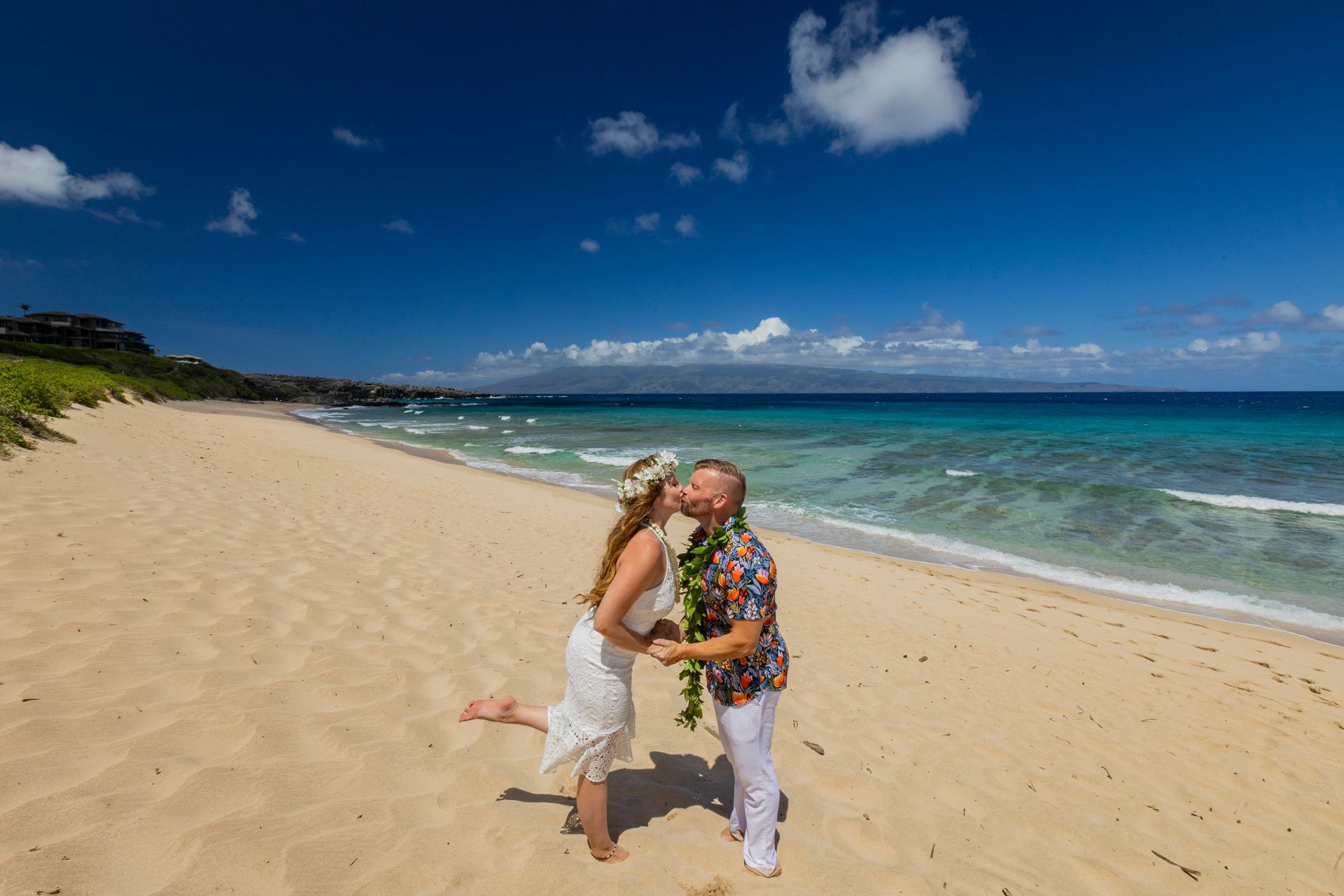 Bride and groom kissing on a sunny beach, blue ocean, white sand. Man wearing leis, woman with flower crown.