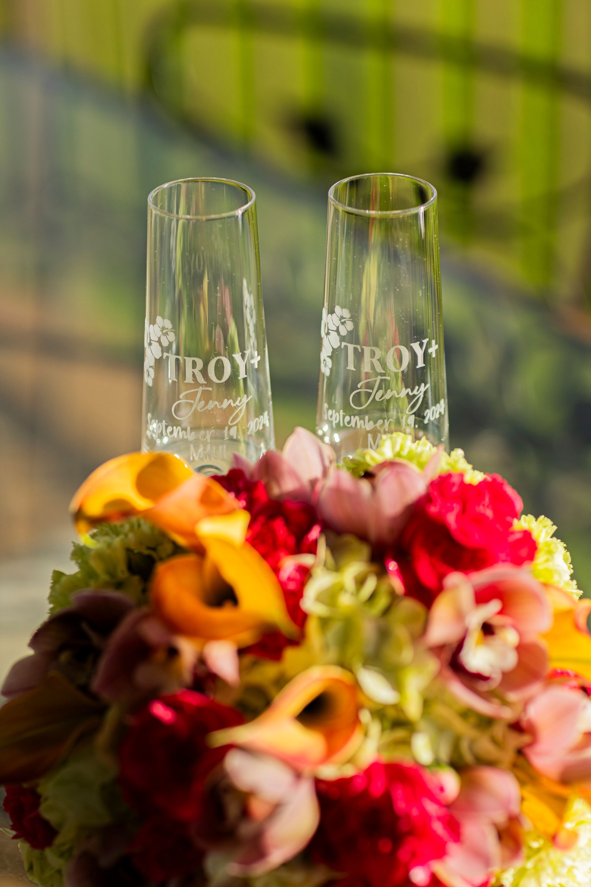 Two engraved champagne flutes atop a floral arrangement, with a blurred background.