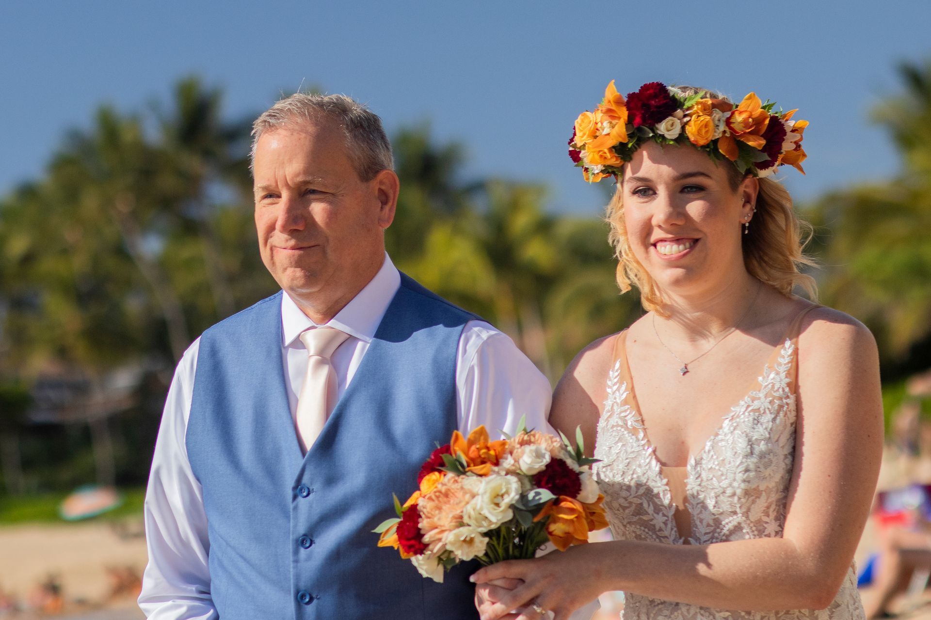 Father walking his daughter down the aisle on a beach. The bride wears a floral crown and holds a bouquet.