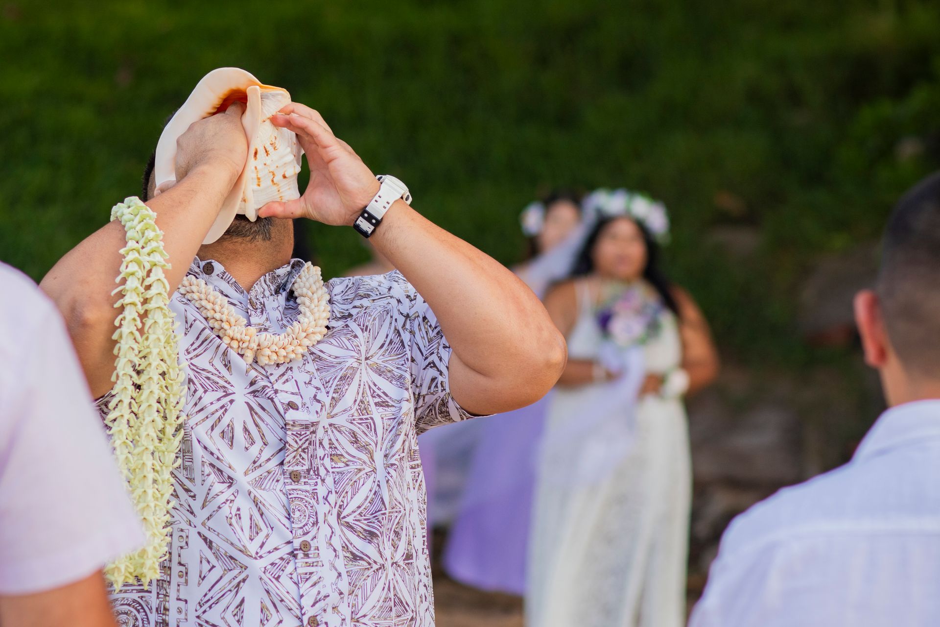 Man blowing a conch shell at a beach wedding; bride and guests in the background.