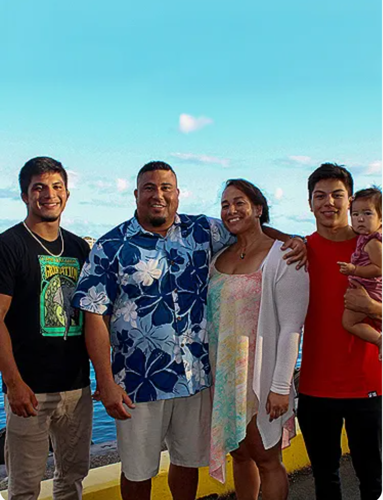 Family of five smiles outdoors by water, under a bright blue sky.
