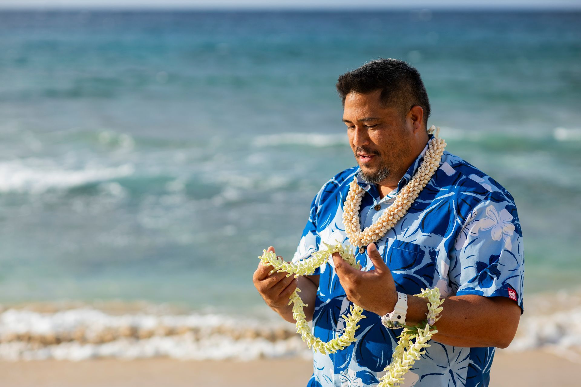 Man on beach holding leis, wearing blue floral shirt. Ocean waves in background.