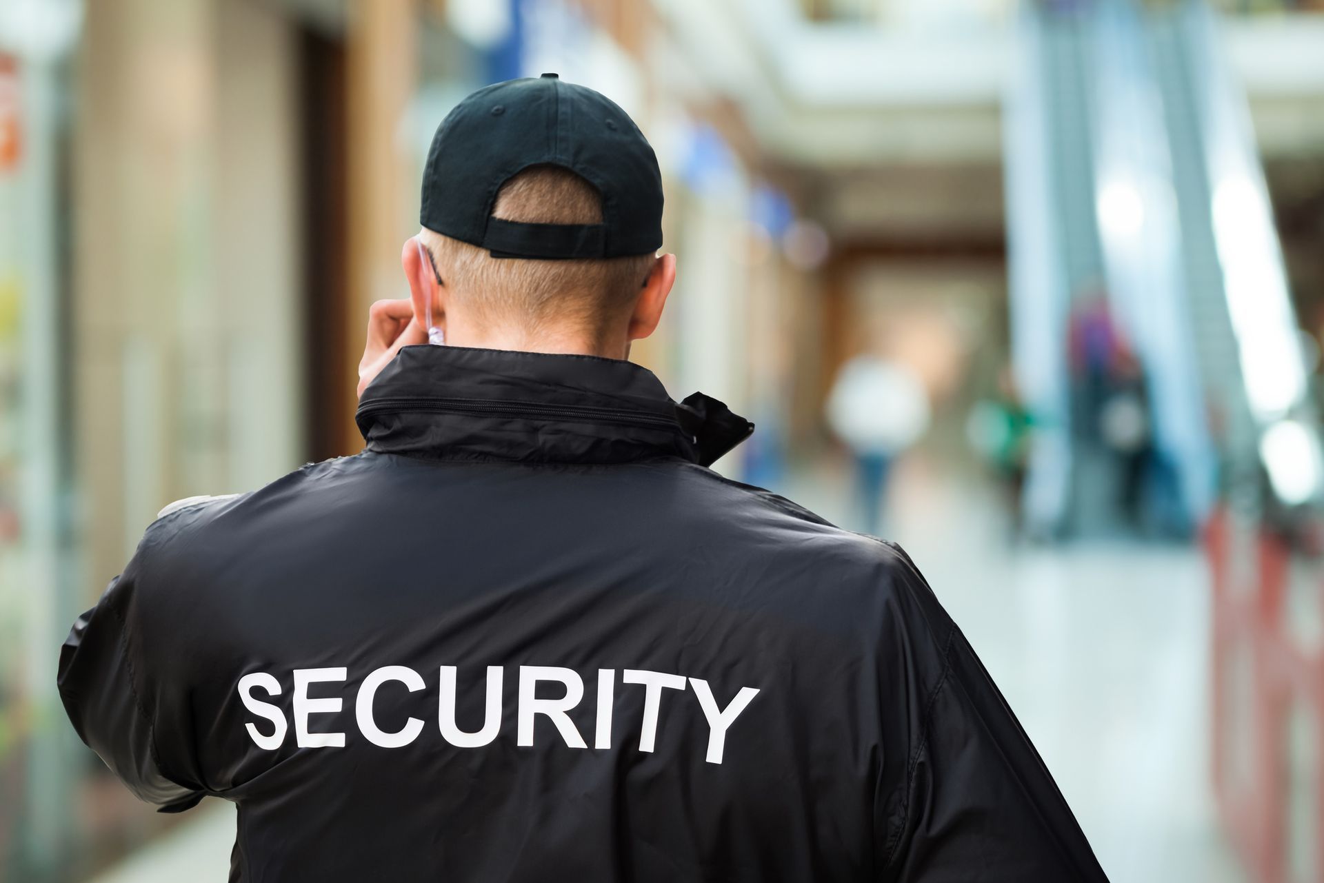 Security guard wearing a black jacket and cap, standing in a shopping mall, looking at blurred background.