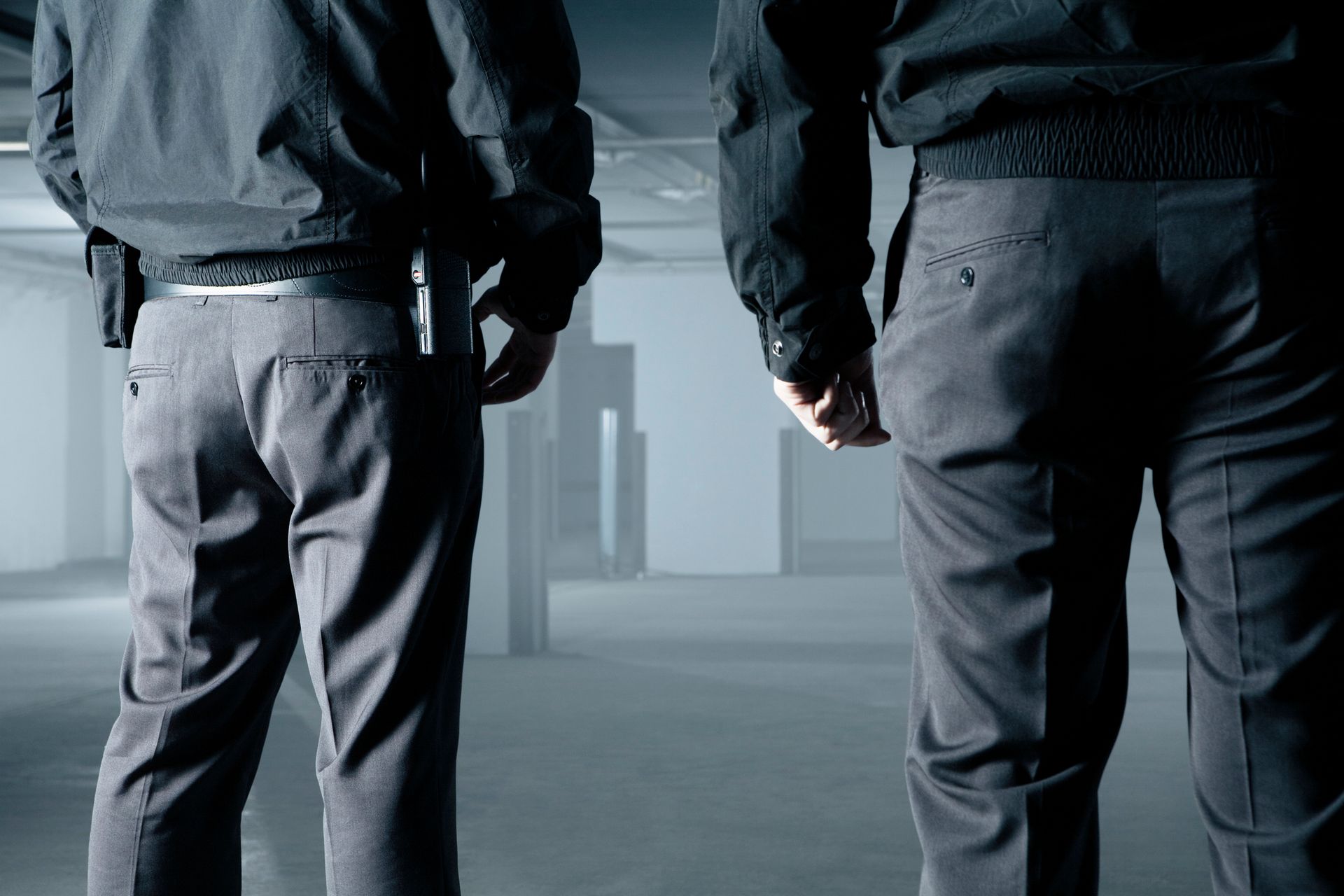 Two figures in dark jackets and pants stand in a large, empty space, facing away.
