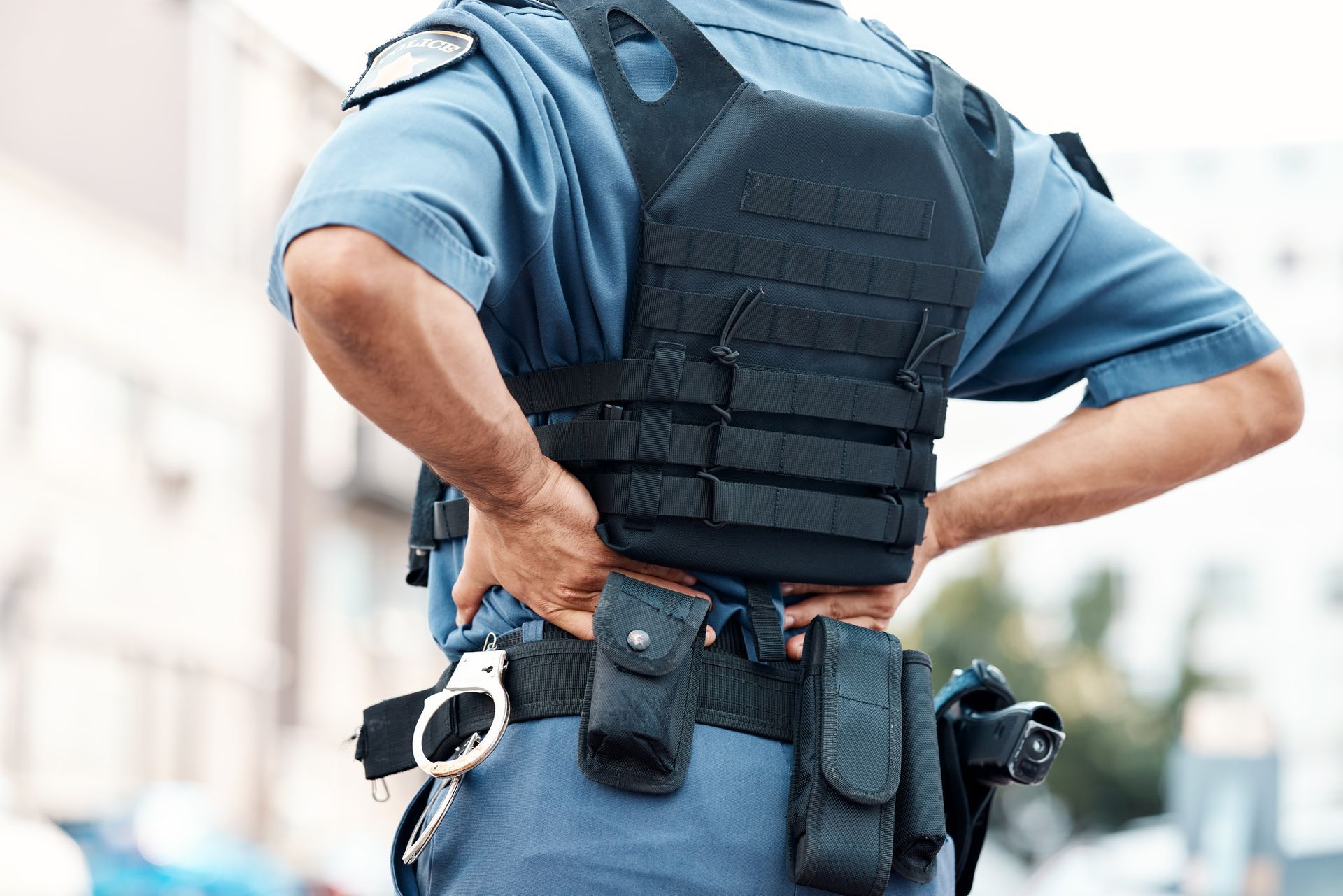 Police officer in uniform, wearing a tactical vest and utility belt, with hands on hips.