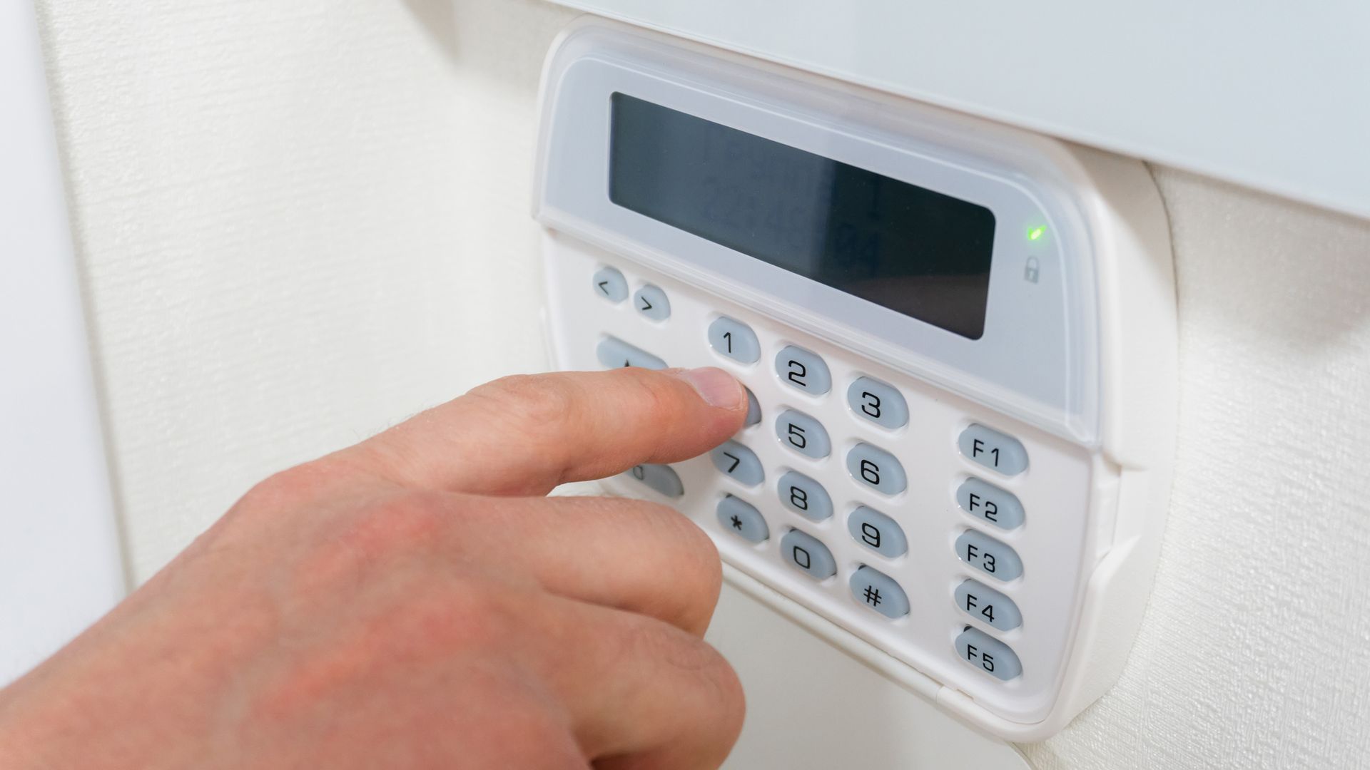 Hand pressing a keypad on a white security alarm panel. Green light lit.