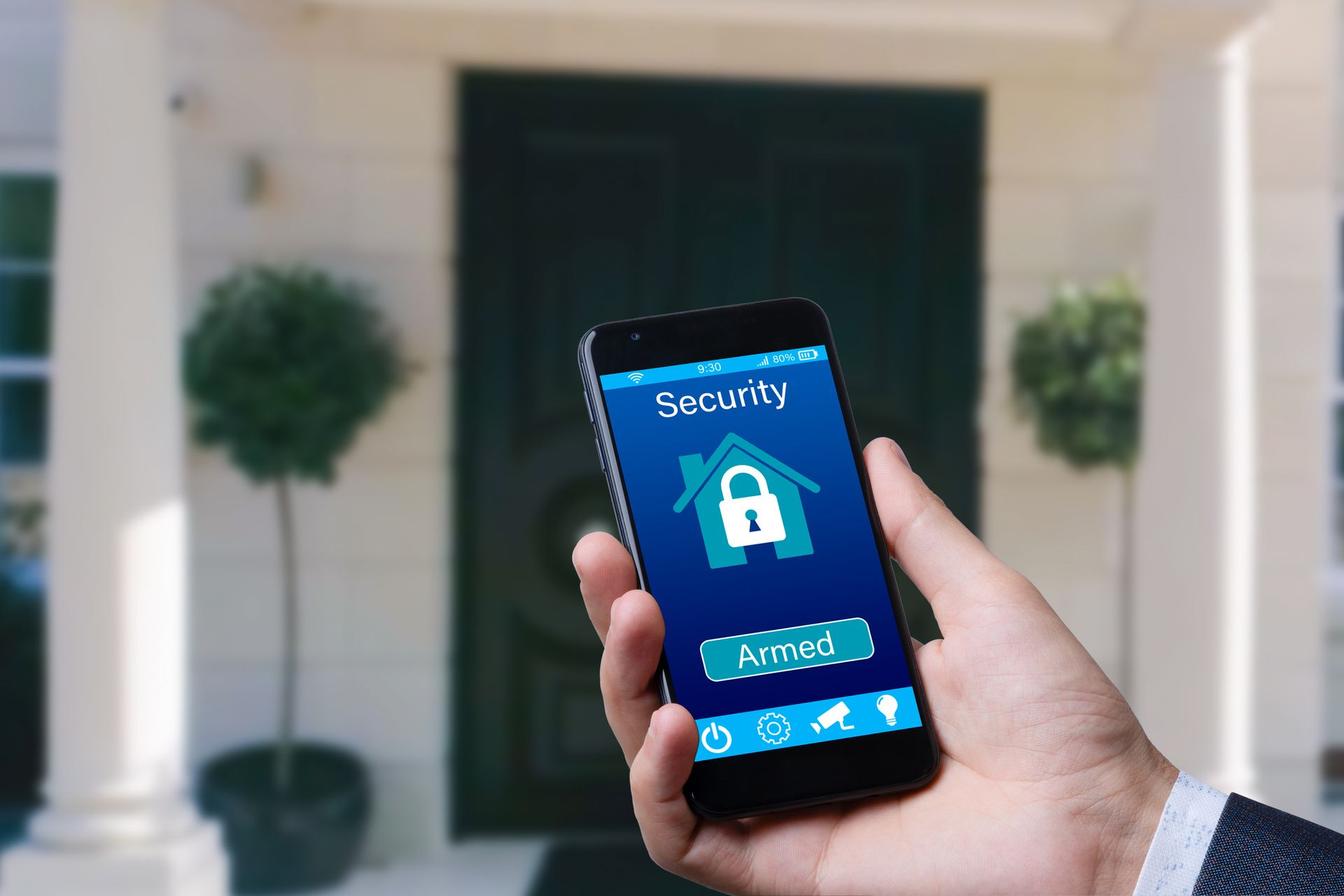 Hand holding a smartphone displaying a security app with a house icon, in front of a house.