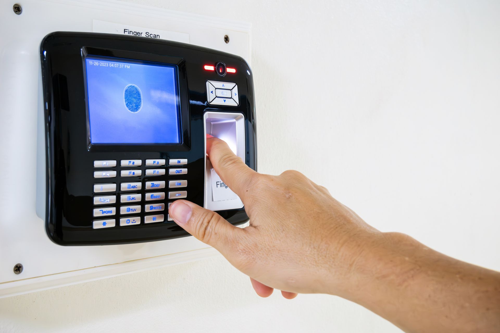 Hand placing finger on a fingerprint scanner, mounted on a white wall.