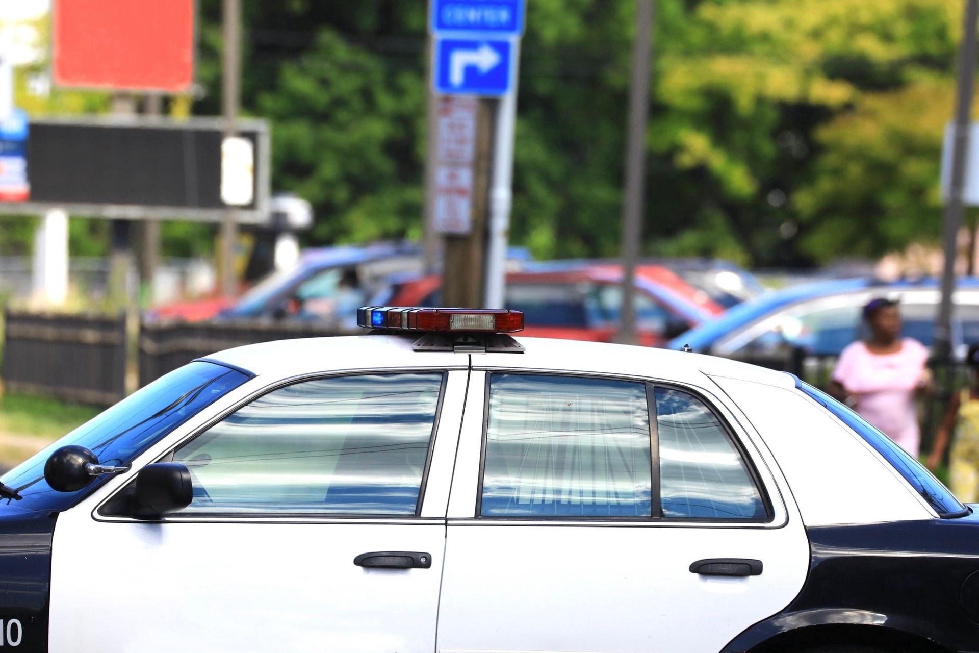 Police car with flashing lights, parked near a street with cars and a person walking in the background.