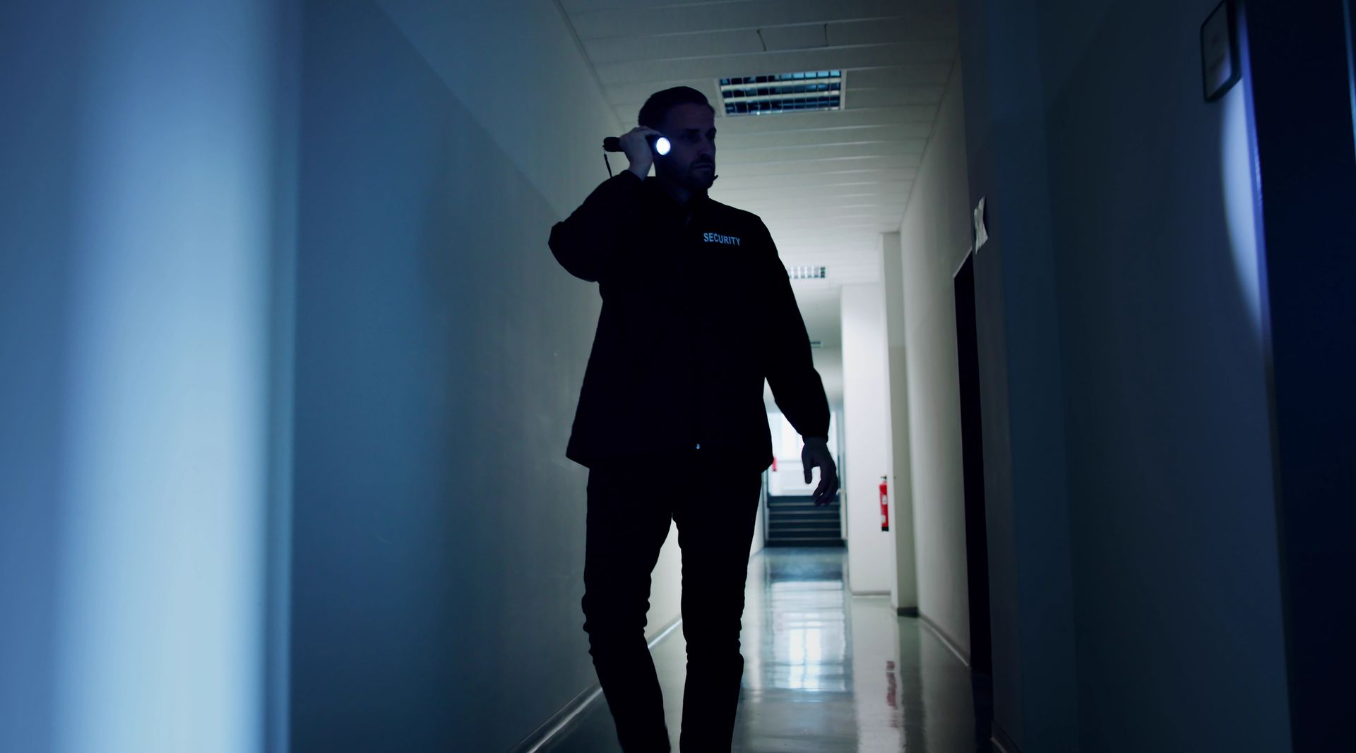 Man with flashlight walks down a dim, narrow hallway.
