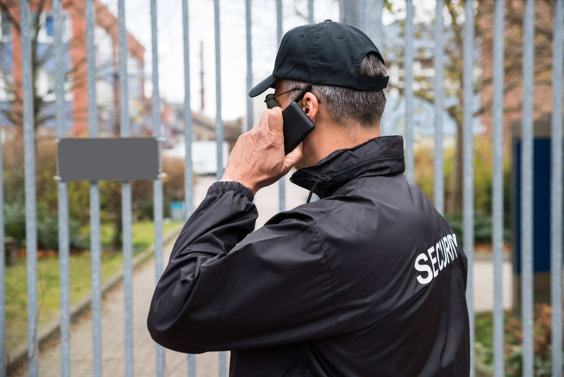 Security guard in black jacket, hat, and glasses talking on phone by a gate.