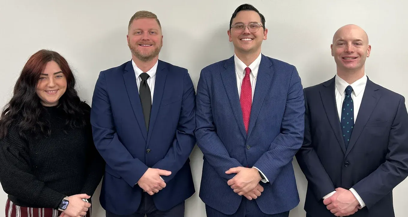 Four people posing for a photo. Three men in suits, one woman in a sweater. White background.