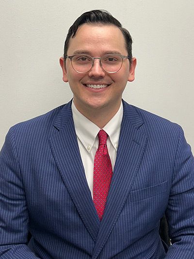 Man in blue pinstripe suit, glasses, and red tie smiling in front of a white background.
