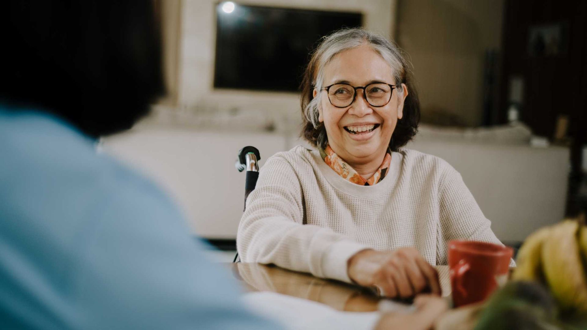 Smiling senior woman in glasses, seated at a table, speaking to a person with their back to the camera.