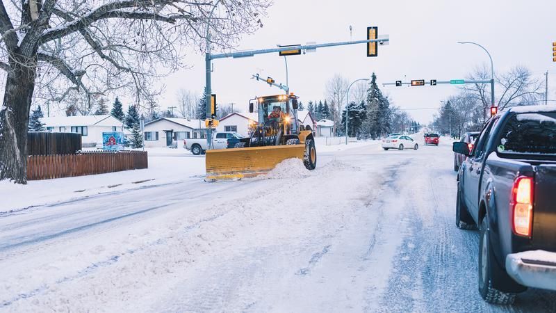 Snowplow clearing a snow-covered road. Cars behind, houses in background, under a snowy sky.