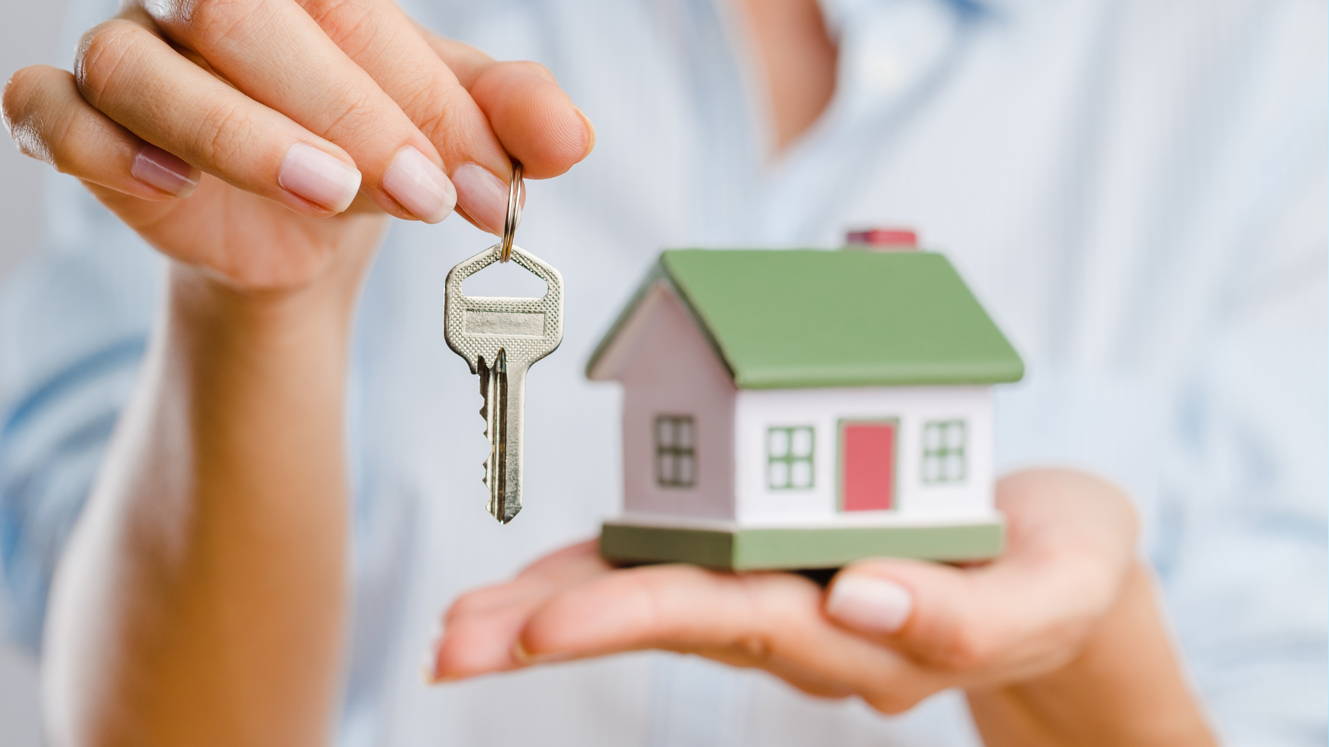 Person holding house key near miniature house.