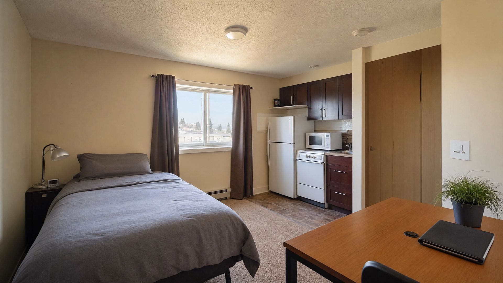 Small bedroom with a bed, dresser, desk, and kitchenette in warm beige tones.