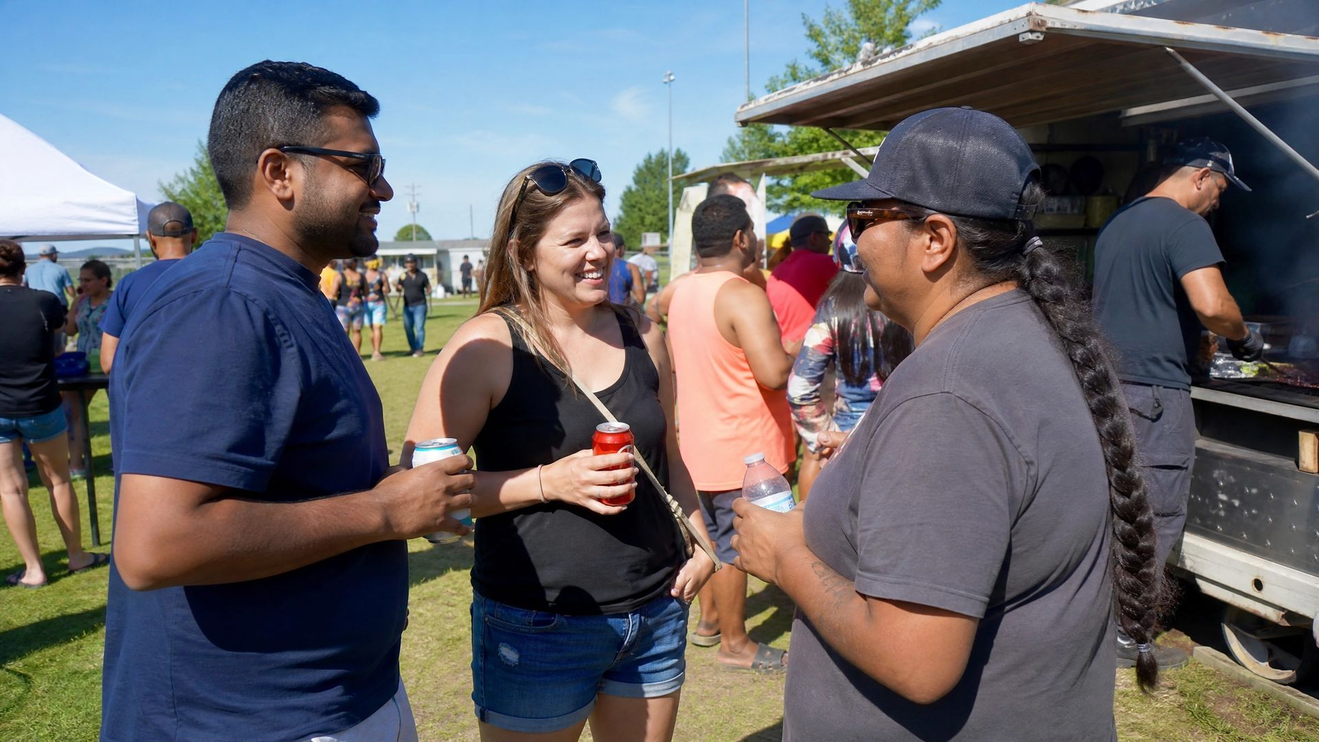 Three people chatting at an outdoor festival, holding drinks near a food stand.