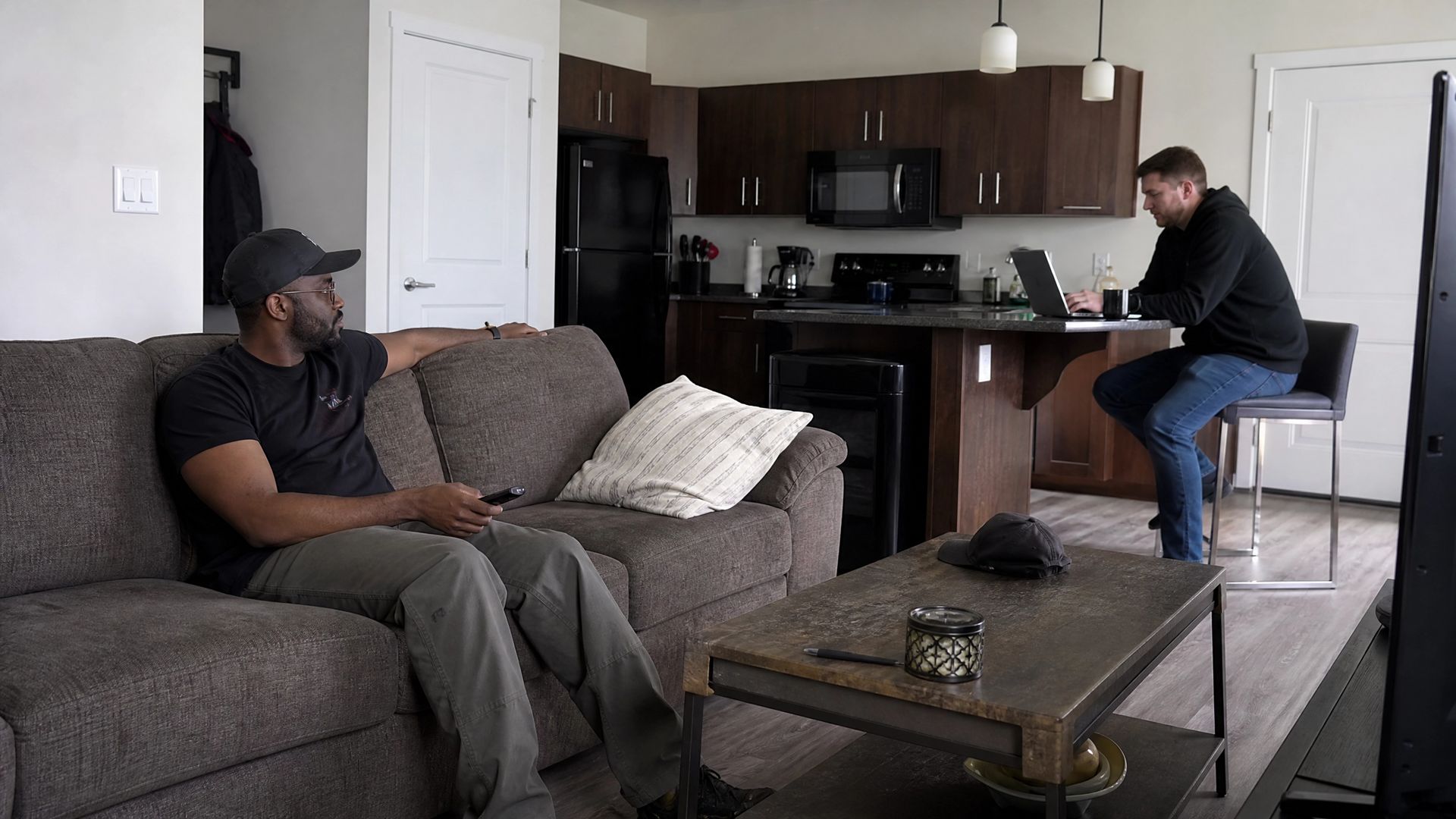 Two people in a living room-kitchen, one on a gray couch and one seated at the counter.