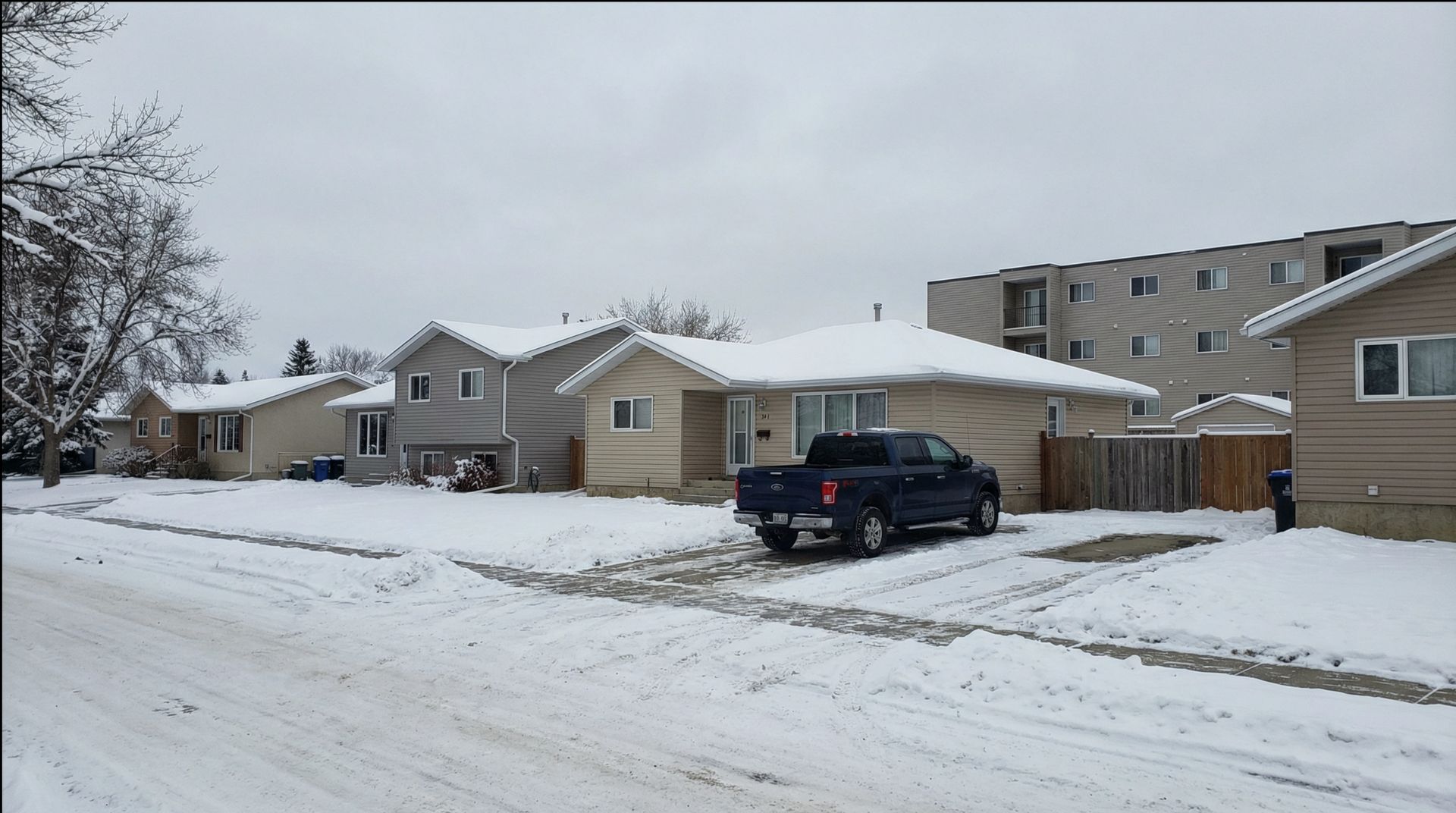 Snow-covered residential street with houses and a truck. Overcast sky.