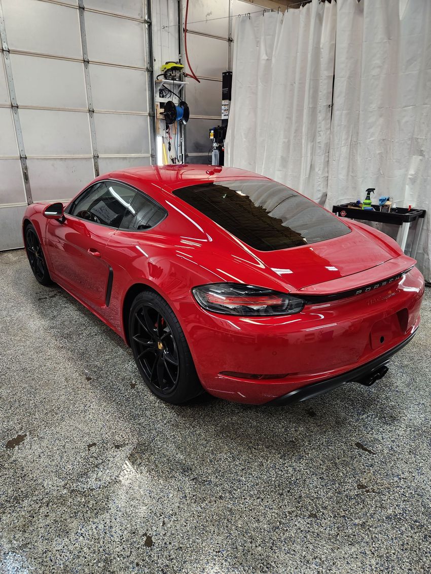 A red Porsche sports car parked inside a garage with a speckled floor and white curtain background.