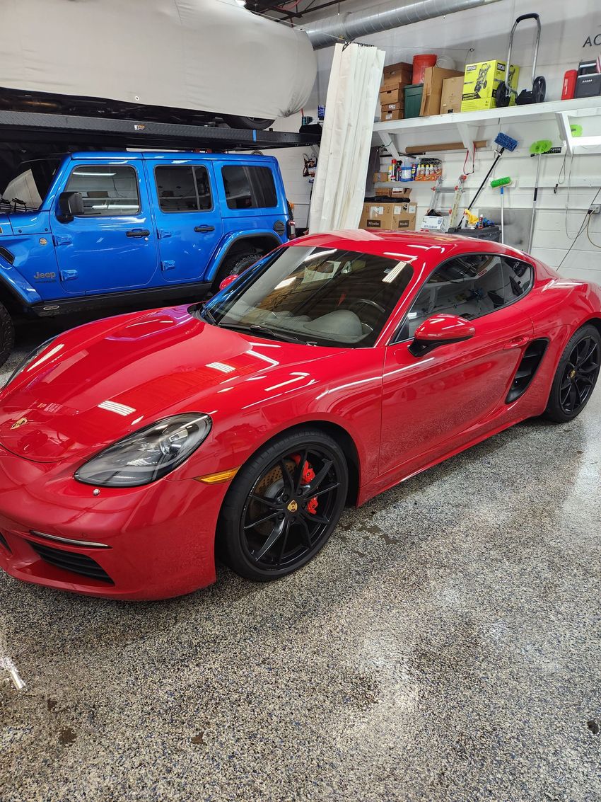 A shiny red Porsche sports car parked in a garage with a blue Jeep visible in the background.