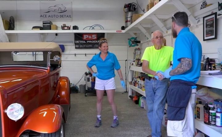 Three people are standing in a garage next to an orange car.