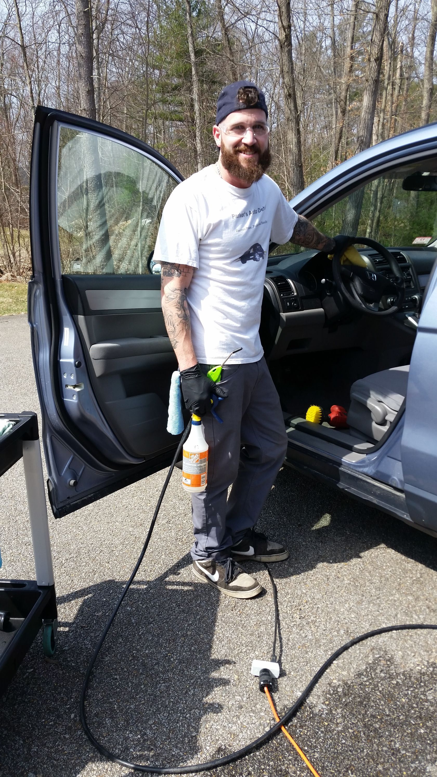 A man with a beard is standing next to a car holding a spray bottle.