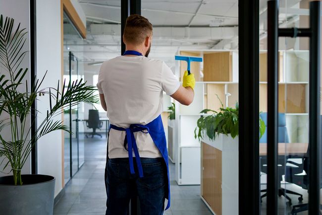 Hombre limpiando la pared de vidrio de la oficina con una escobilla de goma, usando delantal y guantes.