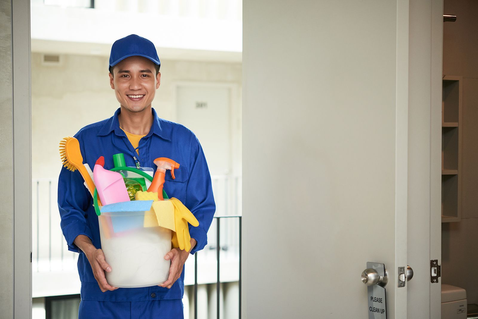 Un hombre con uniforme azul sonríe, sosteniendo un balde con productos de limpieza en una puerta.