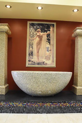 Large stone tub beneath a nude artwork, flanked by ornate pillars, set against a dark red wall.