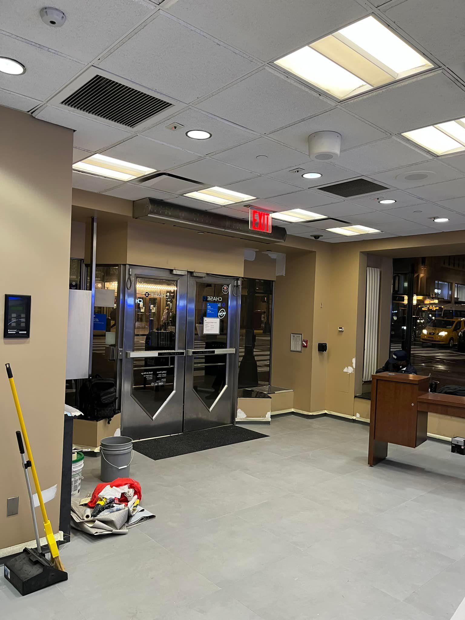 Interior view of a building with an entrance. Beige walls, gray floor, metal doors, and an exit sign are visible.