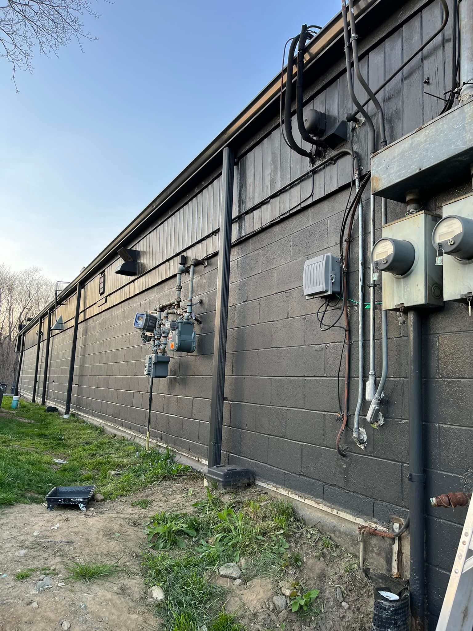 Black building exterior with electrical boxes, pipes, and meters. Green grass in front, blue sky.