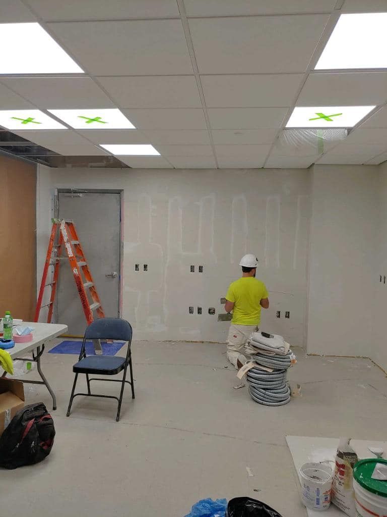 Man in yellow shirt and hard hat spackling a wall in a construction site with a ladder, chair, and materials visible.