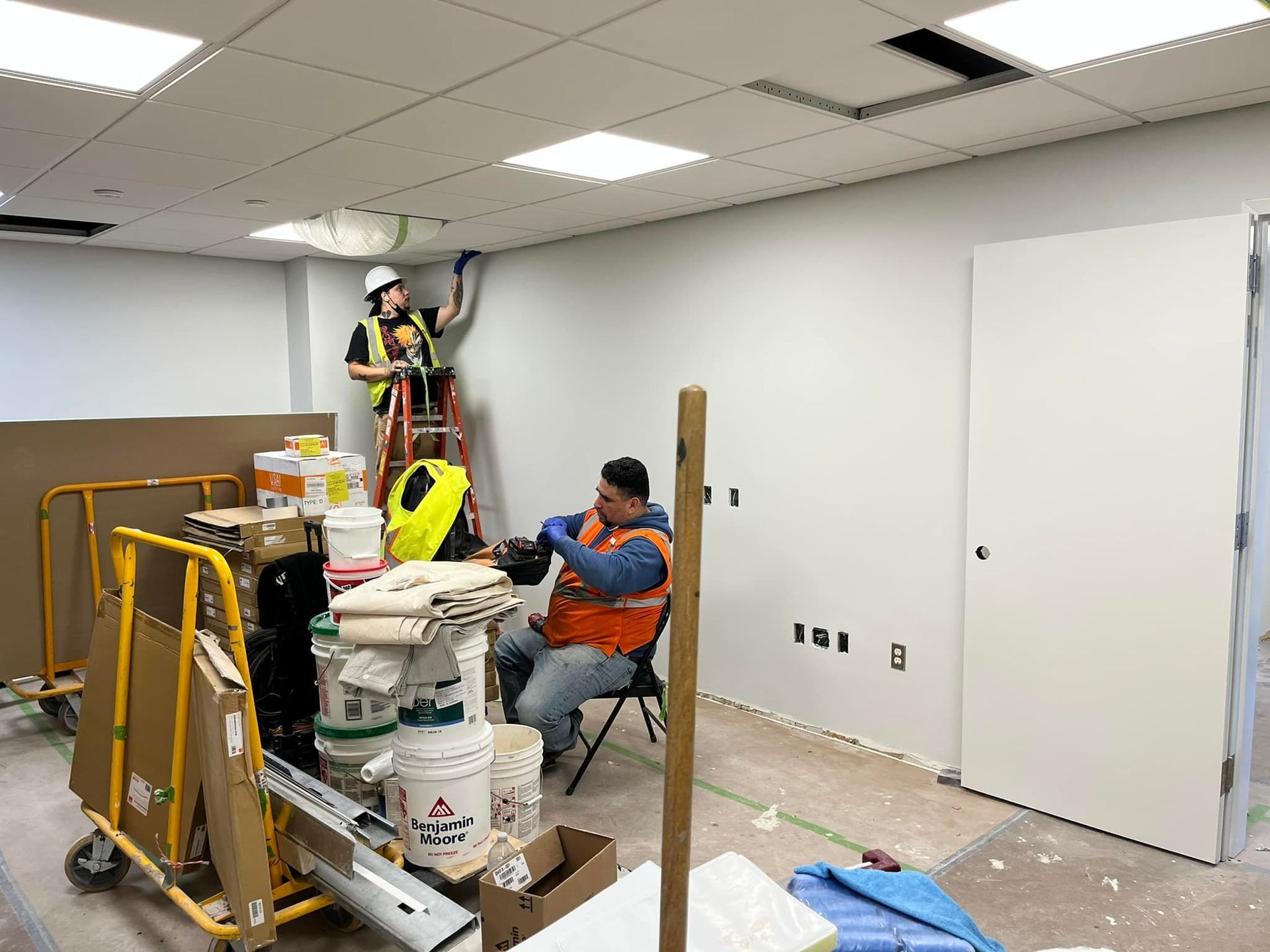 Two construction workers in a room. One on a ladder works on ceiling wiring, the other sits working. The room has materials.