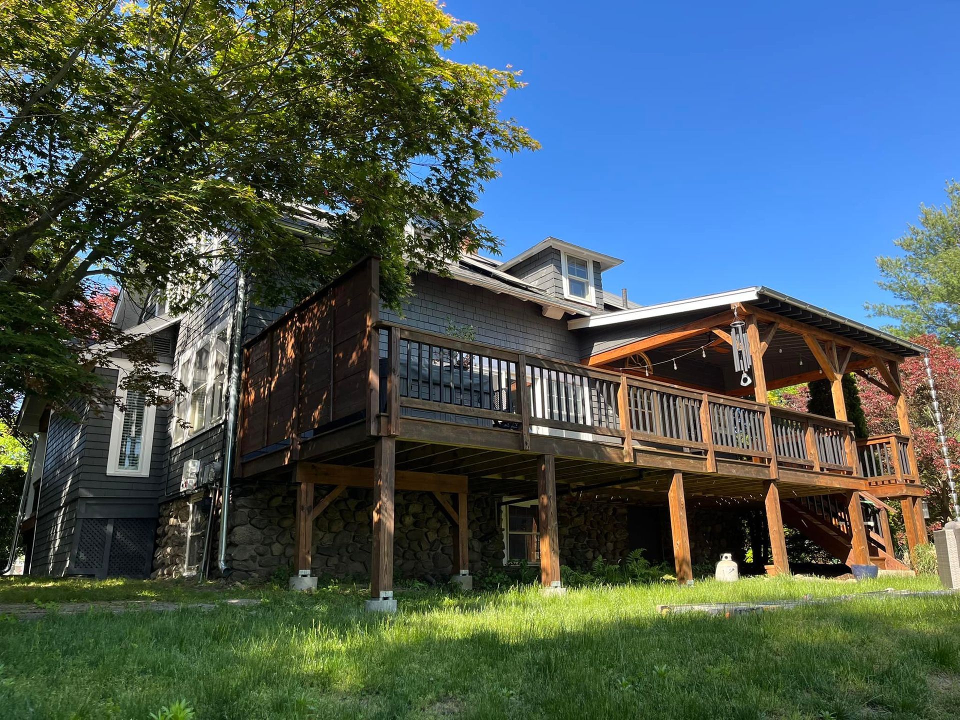 A dark gray house with a wooden deck and a gazebo-style roof in a grassy yard under a blue sky.