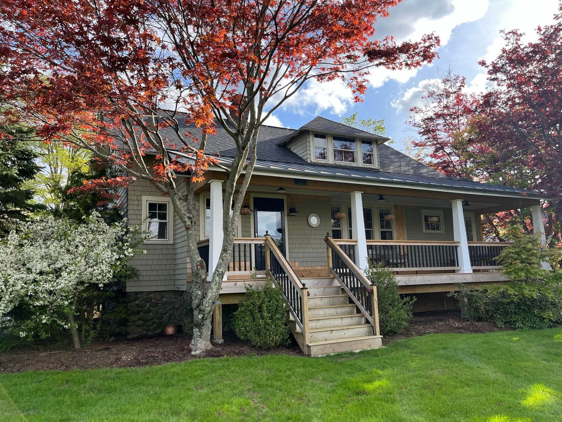 A house with a porch and red-leaved tree. Wooden stairs lead to the front door. Green grass and blue sky.