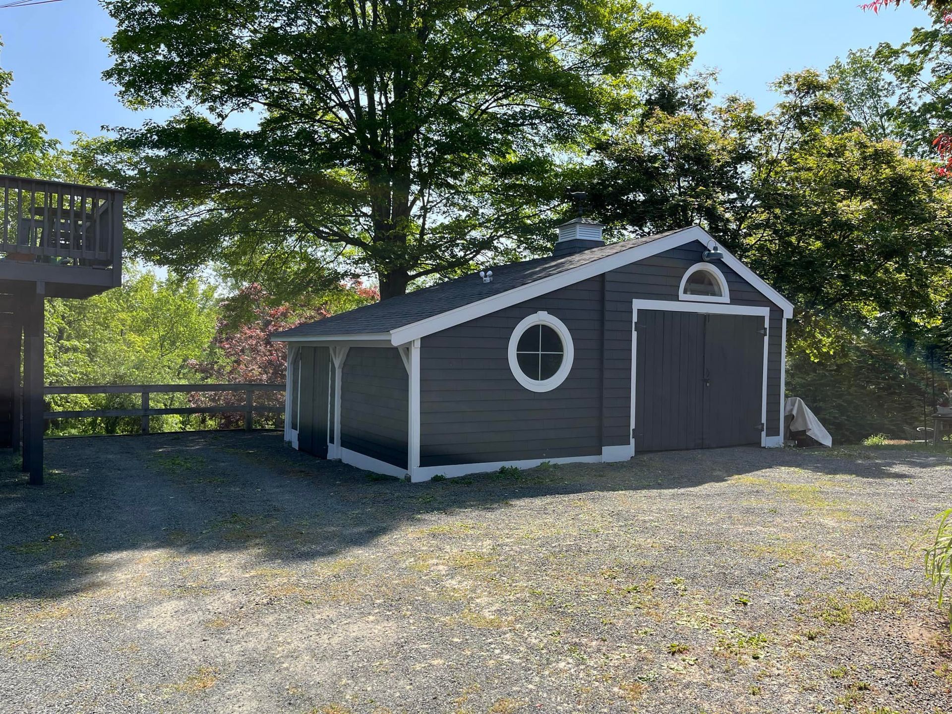 Gray shed with white trim, a round window, and double doors on a gravel driveway, trees in background.