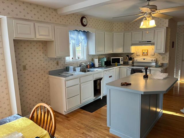 A kitchen with white cabinets, light countertops, an island, a window with curtains, and wallpapered walls.