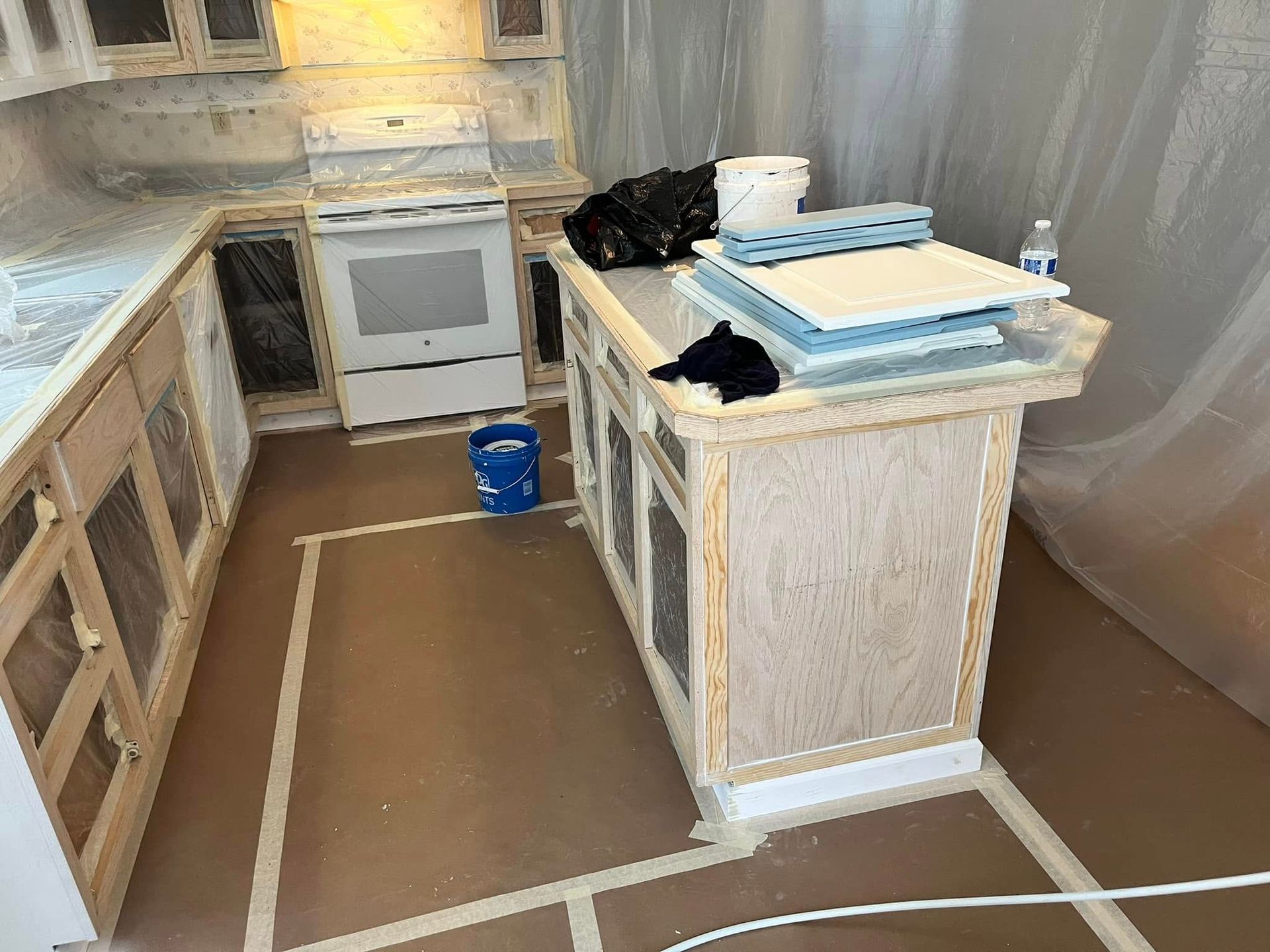Kitchen cabinets and island under renovation; cabinets taped, covered, and supplies on island.