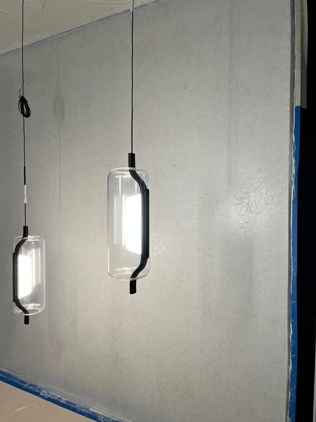 Two modern pendant lights with cylindrical glass shades and black hardware hanging against a plain light gray wall.
