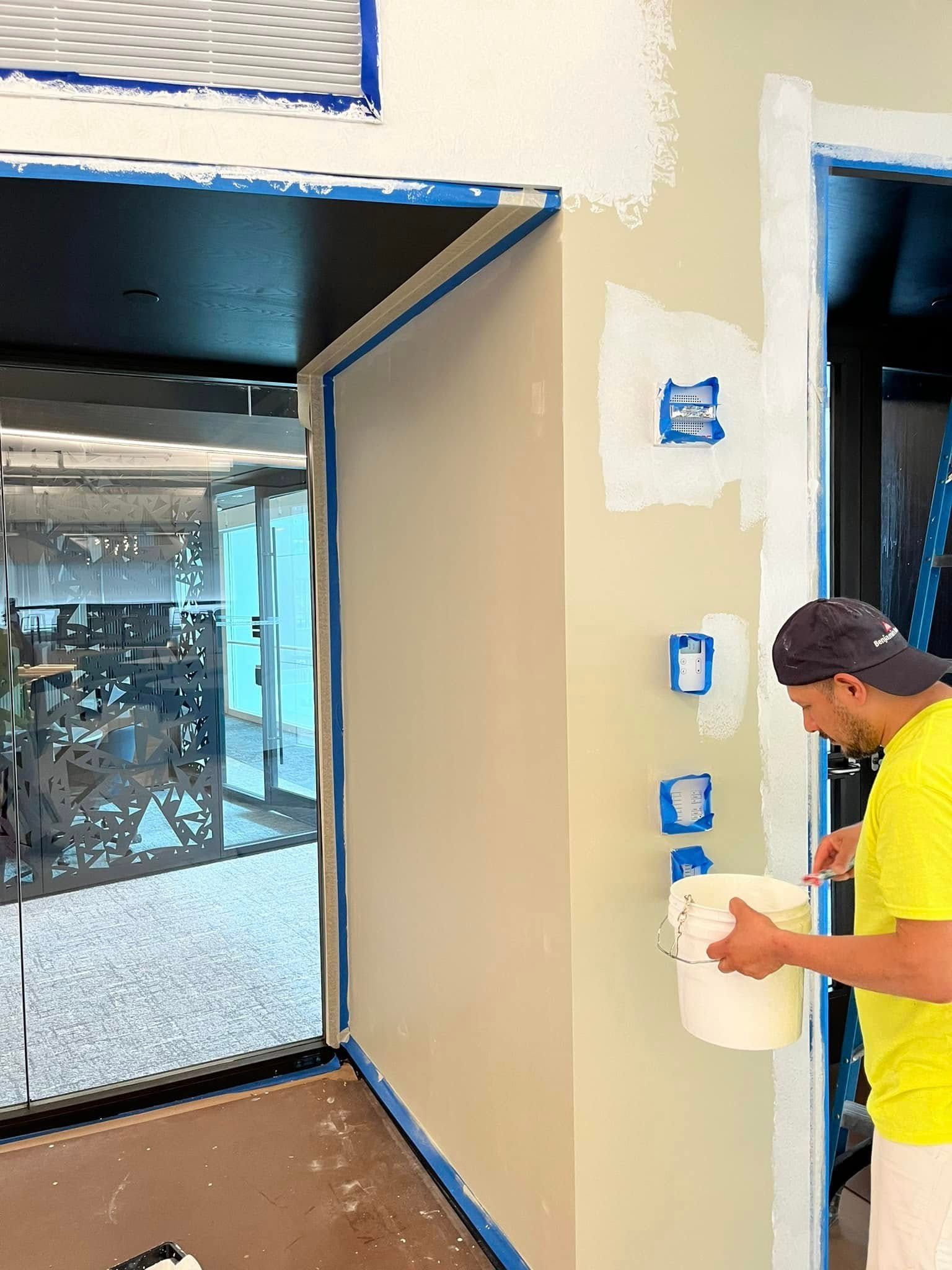 A person paints a wall, interior shot. Blue tape protects edges. White paint. Beige walls, black doorway.