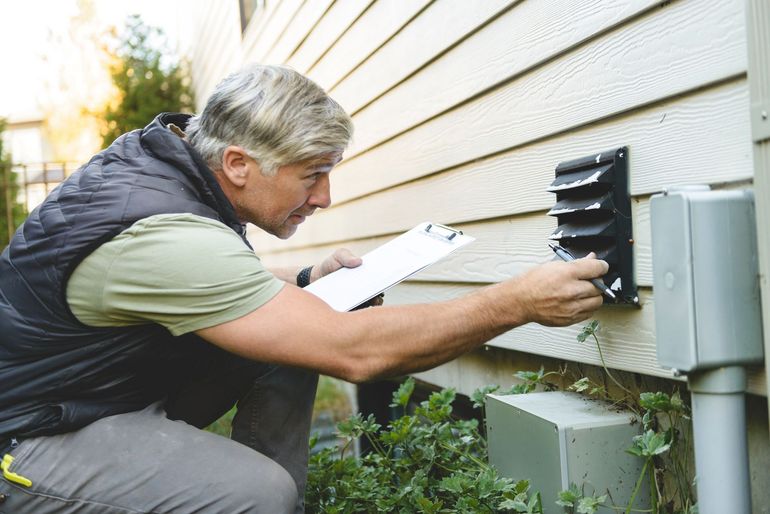 Man examining a wall vent with a clipboard, kneeling outside a building.